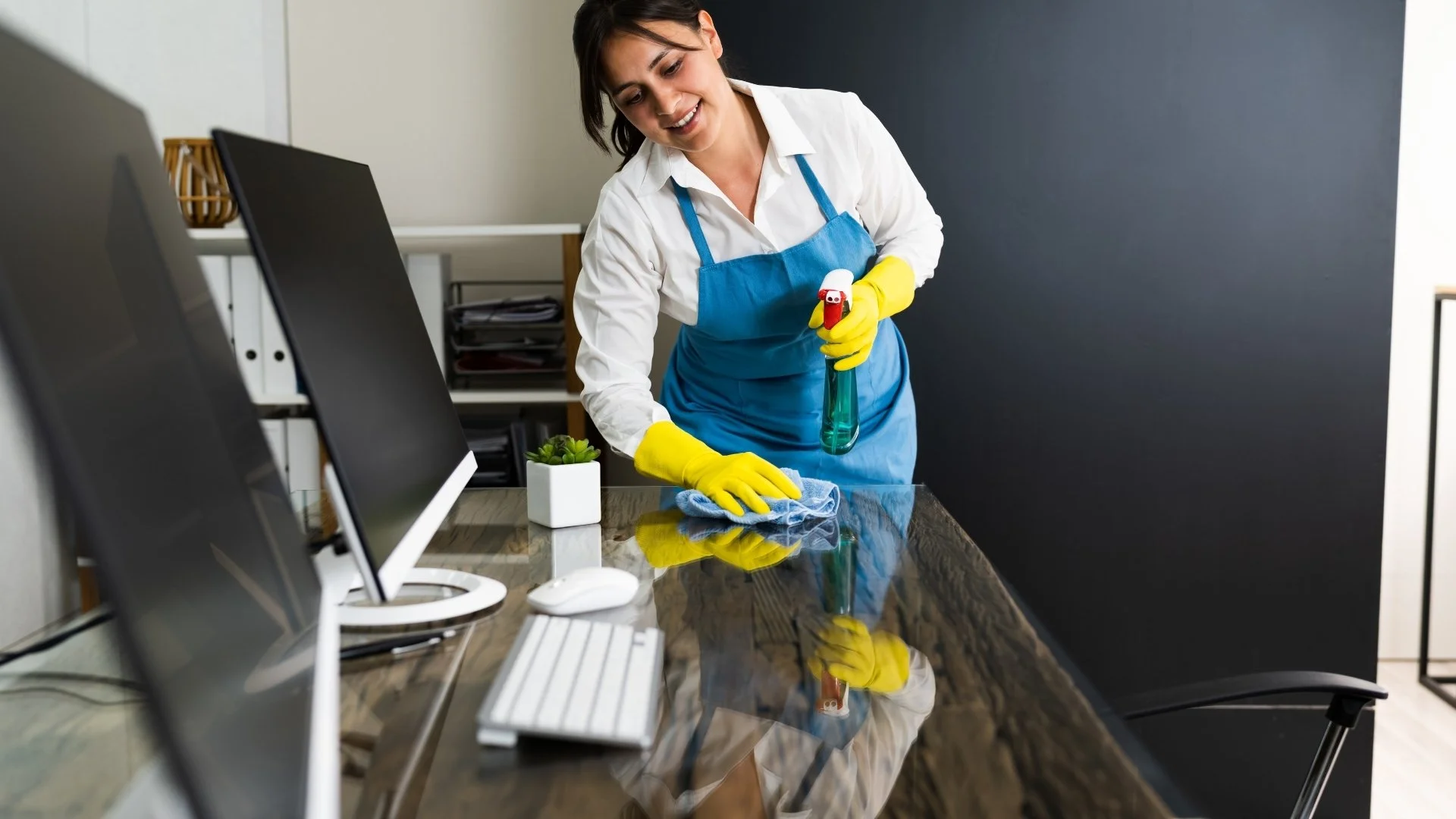 A woman wearing a white shirt, blue apron, and yellow gloves is cleaning a desk with a spray bottle, blue cloth, and wiping cloth in an office.