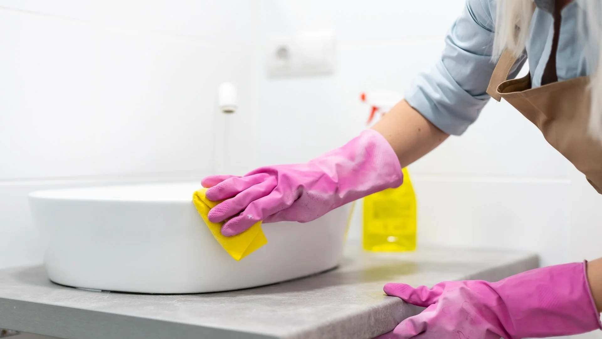 Person cleaning a white bowl with a yellow cloth, wearing pink rubber gloves and a light blue shirt with a beige apron in a clean, white kitchen.