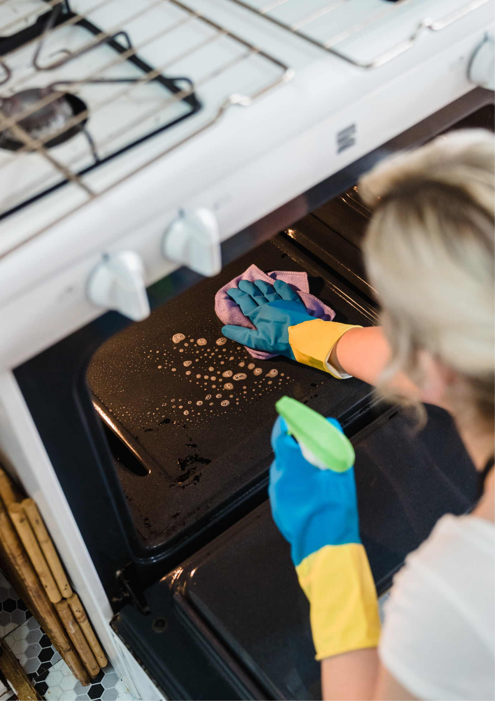 Person cleaning an oven with a pink cloth and wearing blue gloves, seen from above, with part of their blonde hair visible.