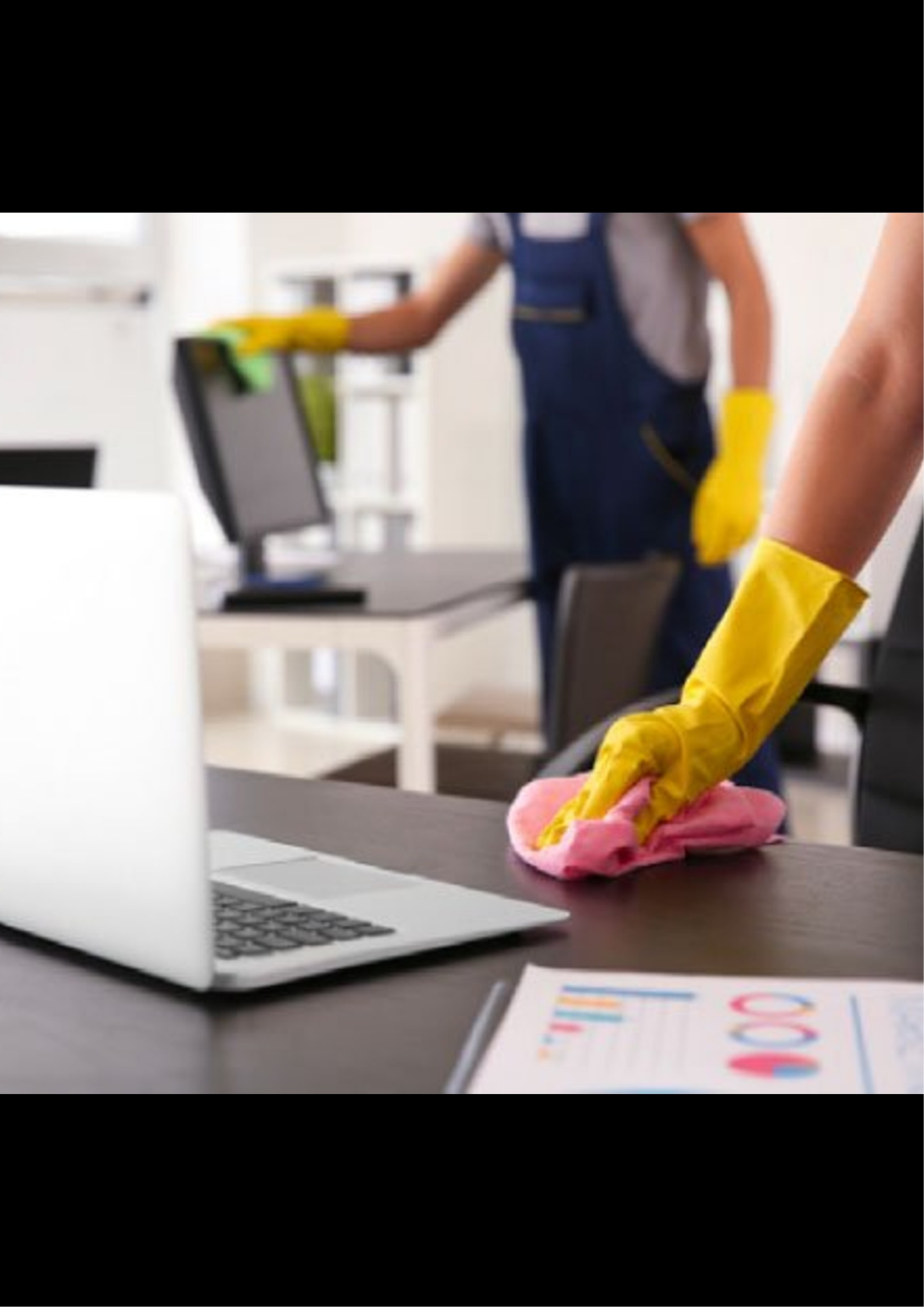 Person wearing yellow rubber gloves cleaning a table with a pink cloth, with an open laptop and papers on the table. In the background, another person is wiping a surface, both dressed in work attire suitable for cleaning or maintenance.