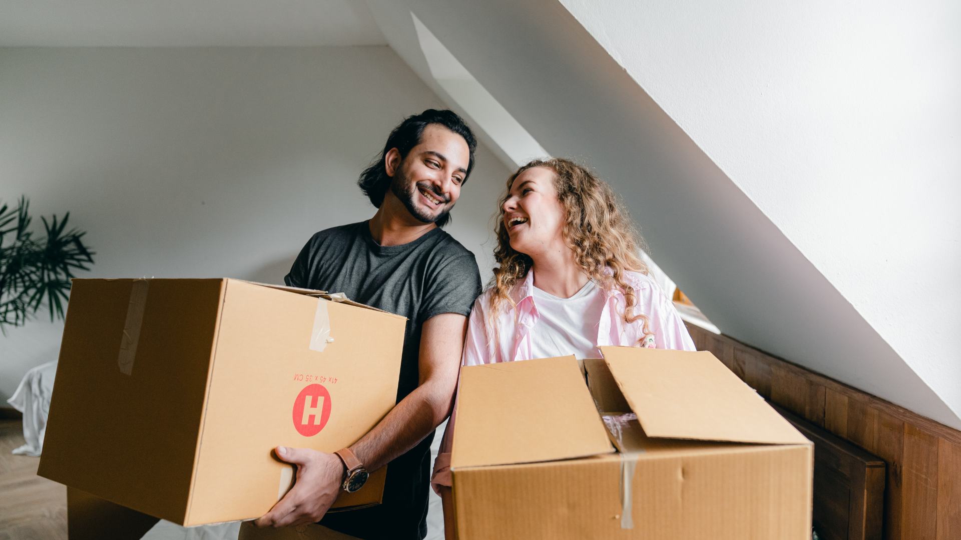 A young man with dark hair and beard, and a young woman with curly blonde hair, are smiling at each other while holding moving boxes in a room with a sloped ceiling and a potted plant in the background.