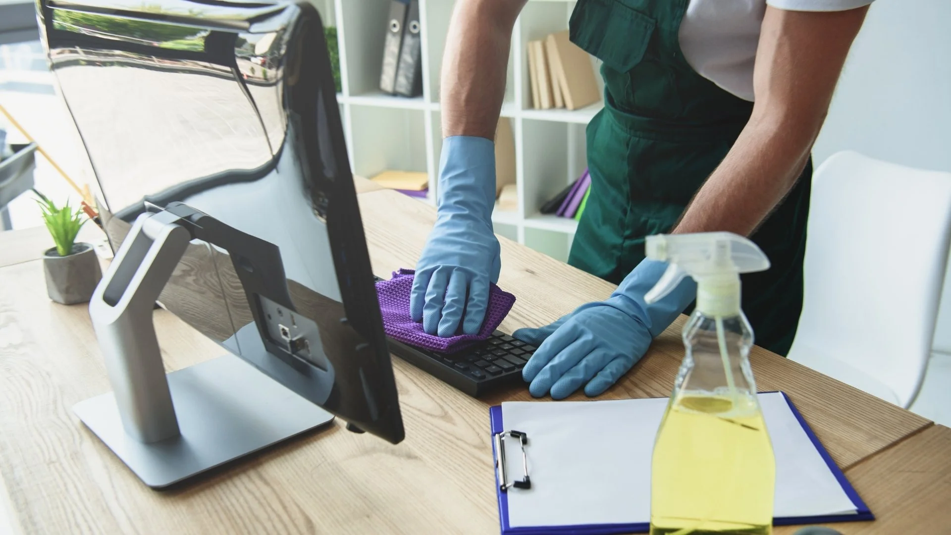Person cleaning a computer keyboard with a purple cloth, wearing blue gloves, in an office setting with a spray bottle of cleaning solution nearby.