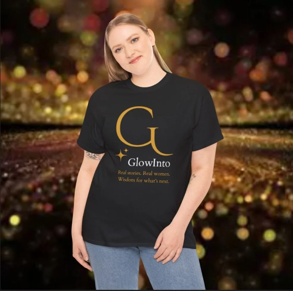 A young woman with blonde hair and tattoos wearing a black T-shirt with gold and white text and design, standing in front of a blurred light-filled background.