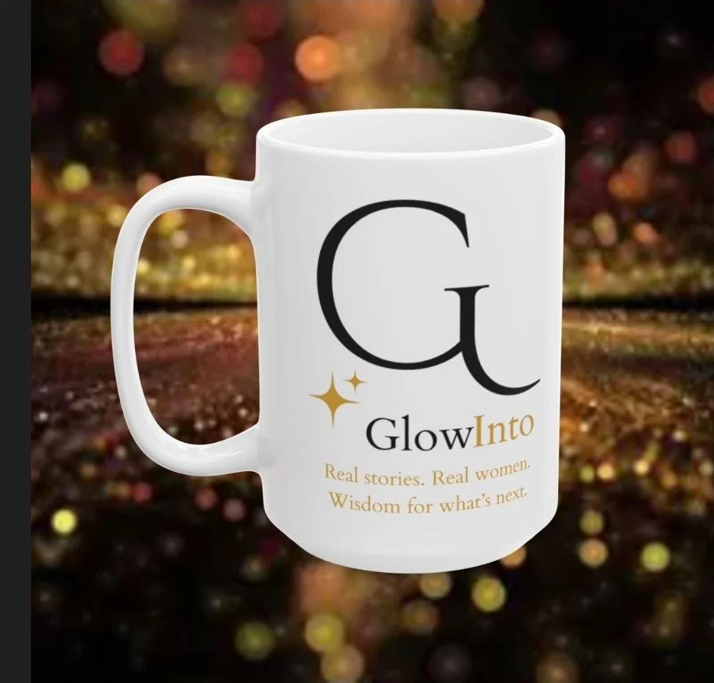 A white mug with the text 'G' and 'Glow Into' printed on it, along with phrases 'Real stories. Real women.' and 'Wisdom for what’s next.' It has a dark, bokeh background with multicolored lights.
