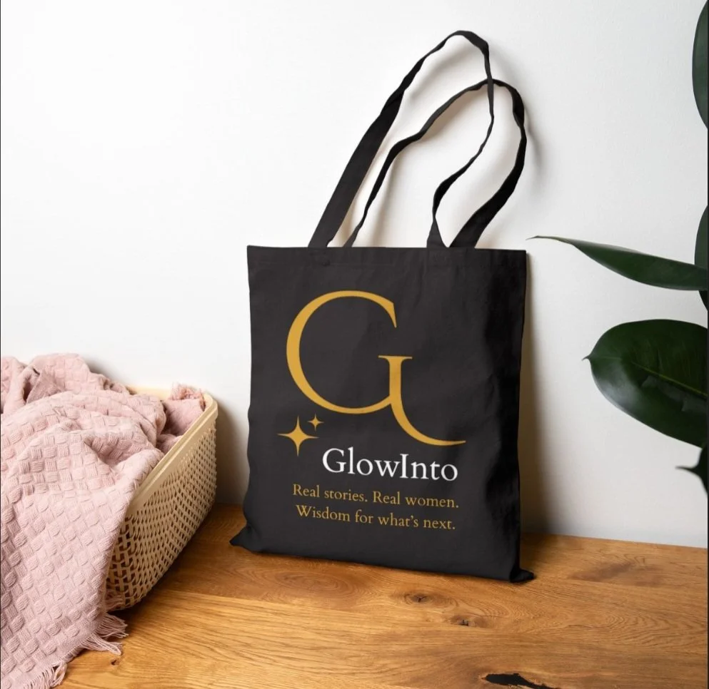 Black tote bag with gold and white text and design, reading "GlowInto: Real stories. Real women. Wisdom for what's next." placed on a wooden surface next to a basket with pink fabric and a green plant.