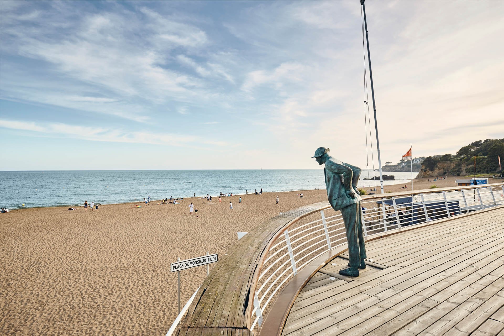 Une plage de sable avec des personnes qui se détendent et jouent près de l'océan. Il y a une statue en bronze d'un homme portant un chapeau, penché en avant, située sur une promenade en bois avec une rampe blanche. Le ciel est partiellement nuageux et il y a des falaises avec des végétations en arrière-plan.