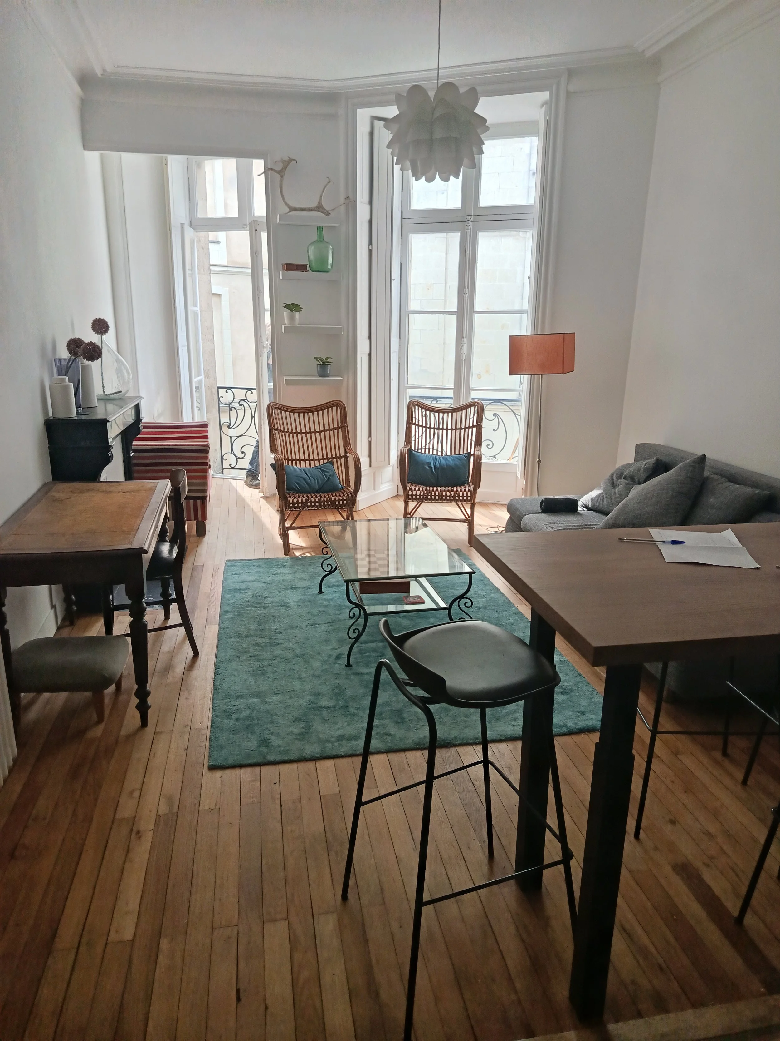 Salon avec deux fauteuils en bois, canapé gris, tapis vert, table basse en verre, grande fenêtre laissant entrer la lumière, étagère avec des plantes et des objets décoratifs, lampe orange, bureau en bois avec papier et stylo.