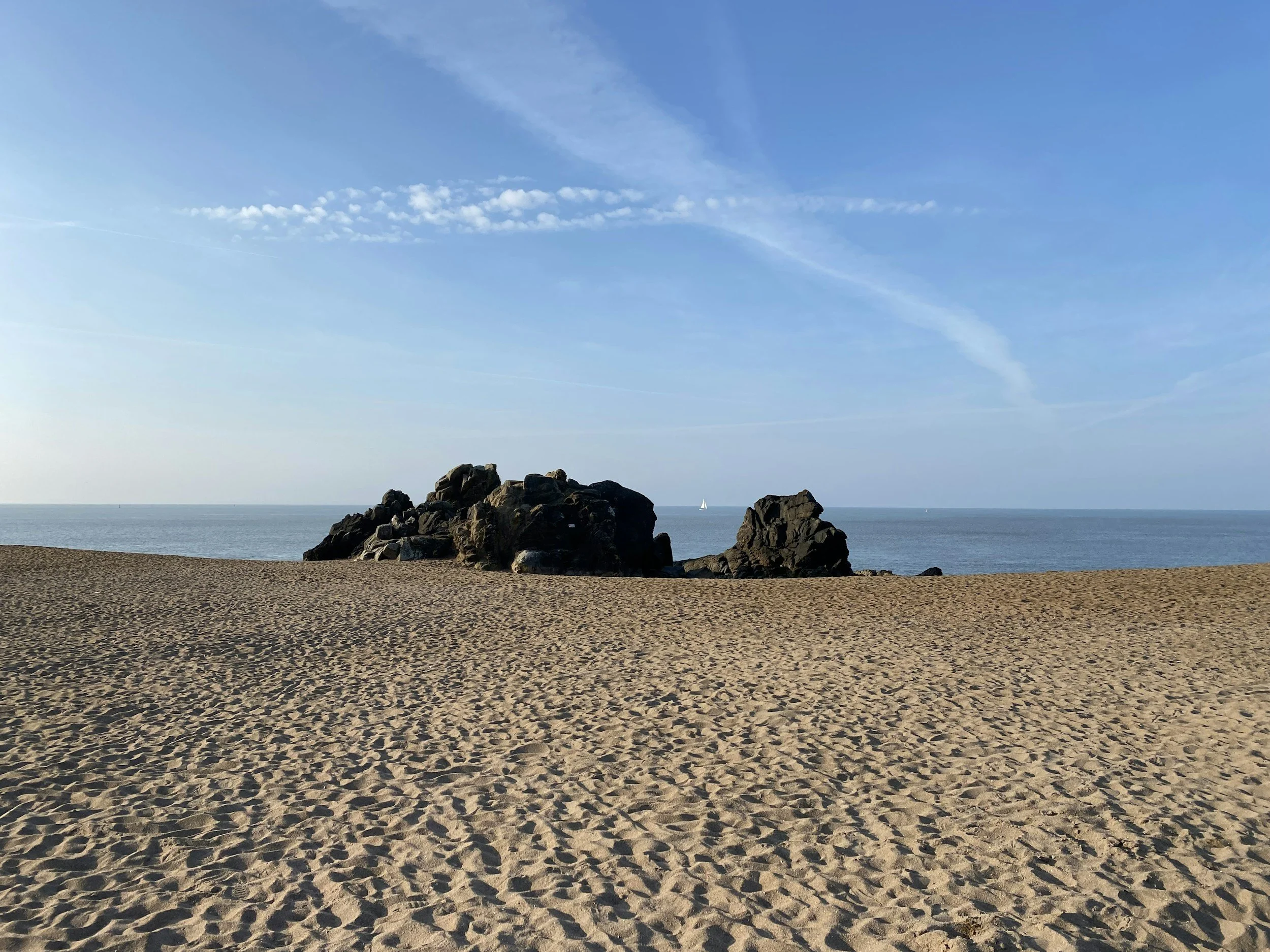 Plage de sable avec des rochers au centre et un bateau à voile en arrière-plan, ciel bleu avec quelques nuages.