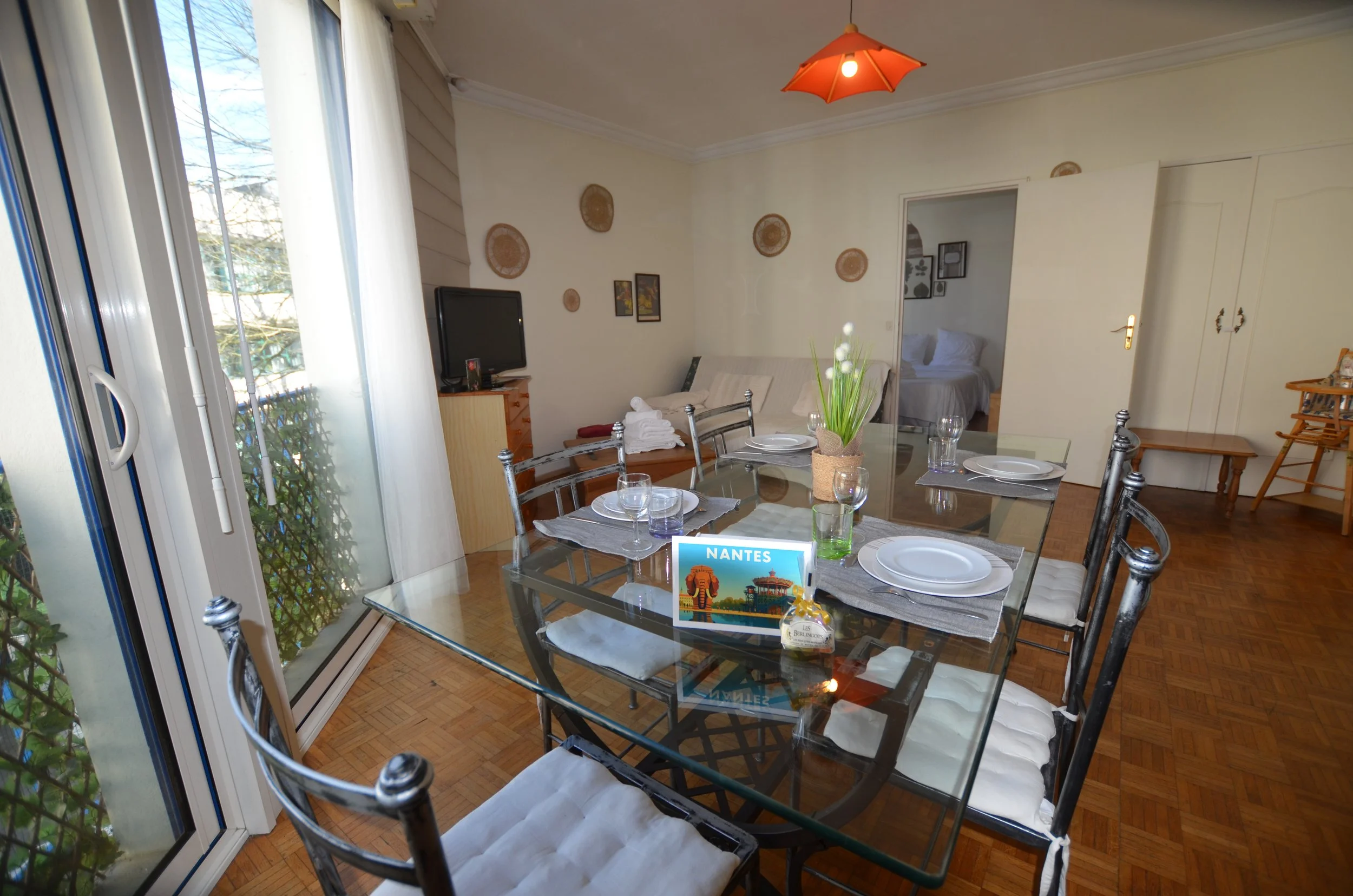 Salle à manger avec table en verre, assiettes, verres, coussins blancs sur les chaises, décoration murale, télévision, porte menant à une chambre, fenêtre avec vue sur le jardin, lampe suspendue rouge en forme d'ombrelle.
