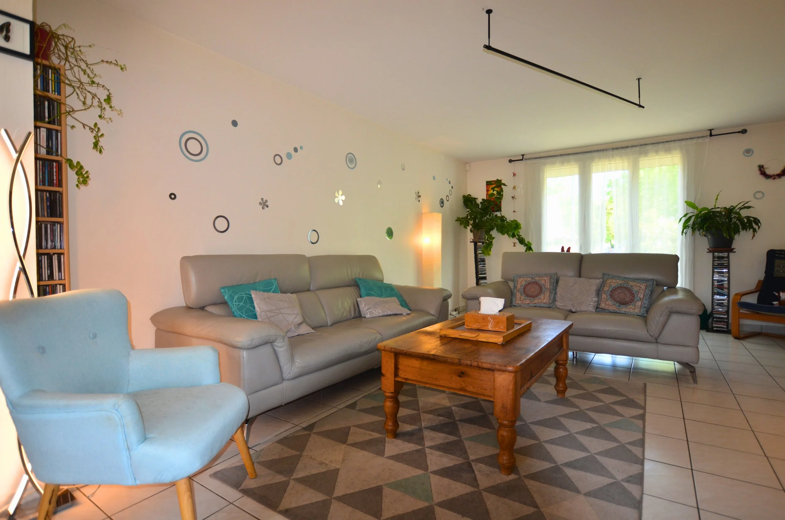 Salons avec canapé en cuir beige, plantes vertes, meuble en bois, mur décoré avec cercles et fleurs, grande fenêtre avec rideaux blancs, sol en carreaux, éclairage naturel.