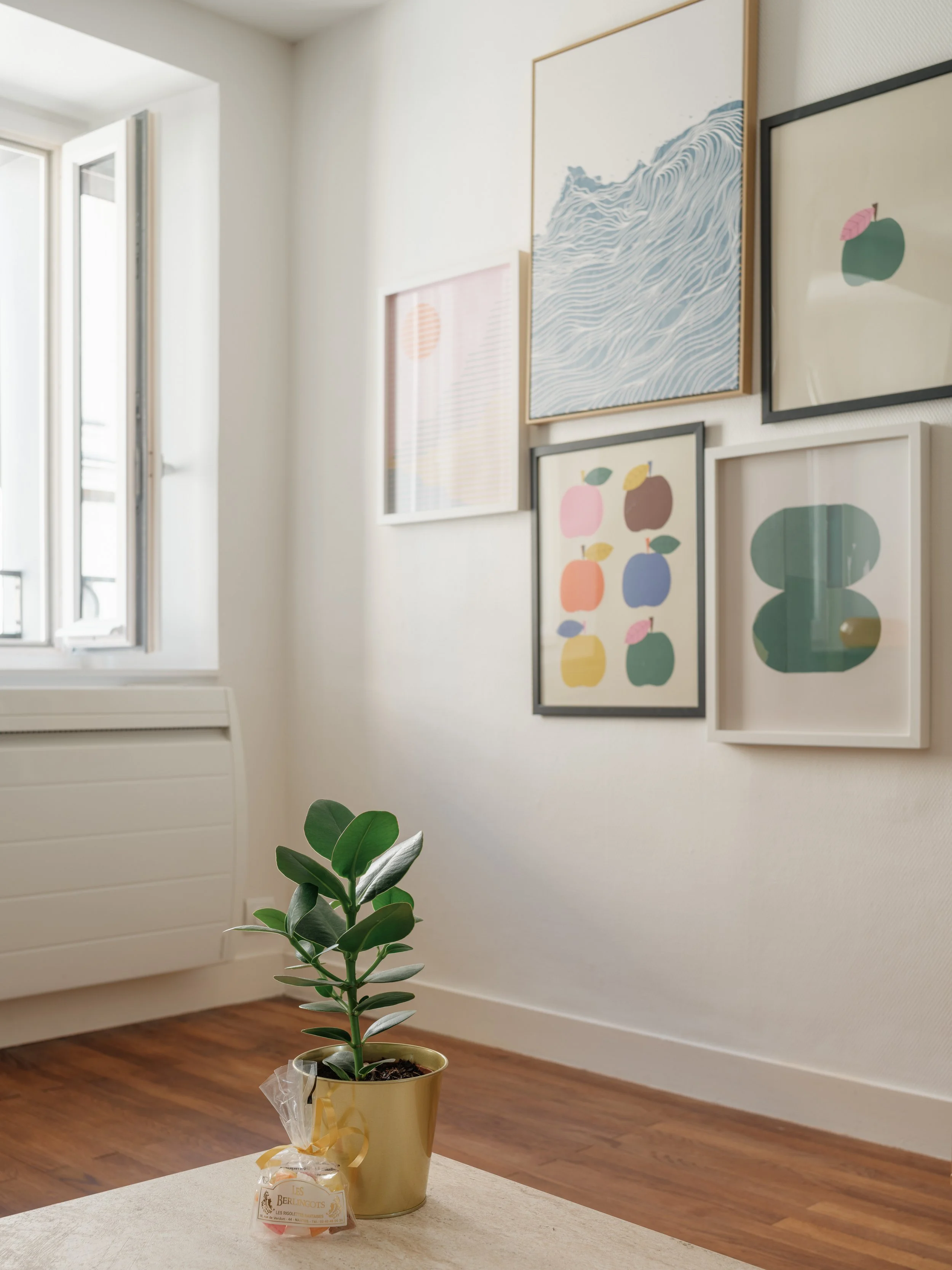 Une plante verte en pot doré posée sur une table avec un petit sachet de bonbons à côté. En arrière-plan, une murale avec des œuvres d'art modernes et un mur blanc avec une fenêtre ouverte.