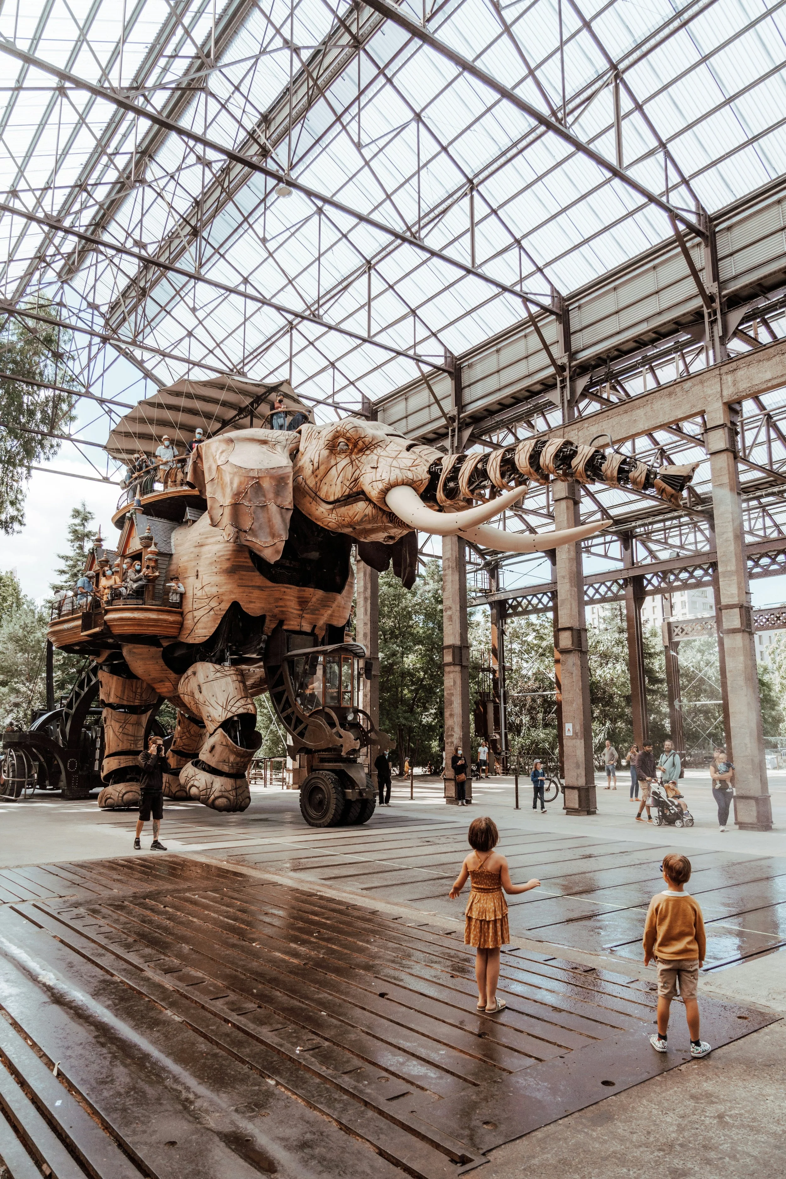Une grande sculpture d'un éléphant en bois située dans un espace couvert avec une structure en métal et en verre. Des enfants et des adultes interagissent autour de l'œuvre, qui semble présenter un habitat de forêt tropicale sur son dos.