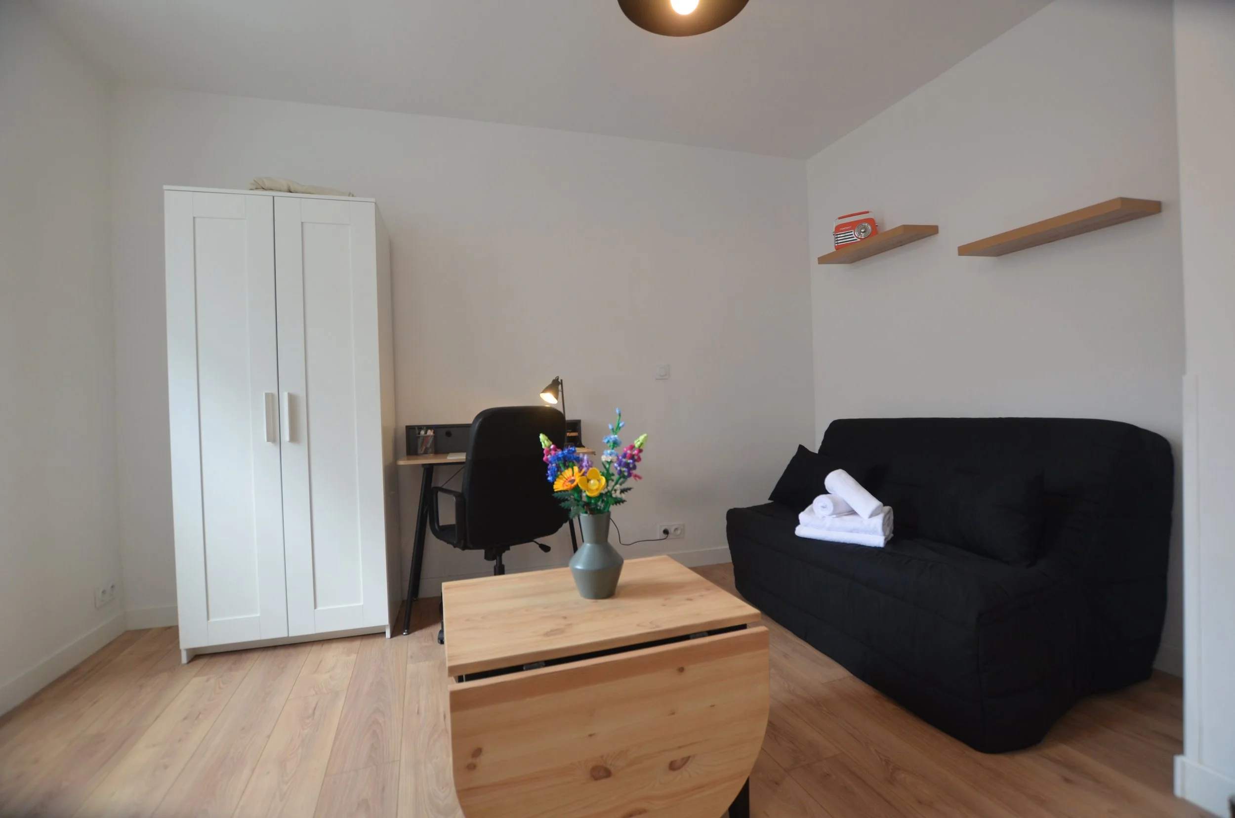 Salon avec un canapé noir, une table en bois, une lampe de bureau, des étagères en bois, une vase avec des fleurs colorées, et des serviettes blanches sur le canapé.