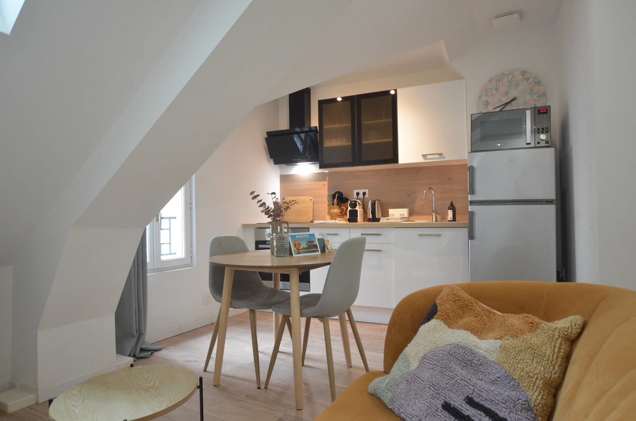Intérieur d'une cuisine moderne dans un appartement lumineux avec un coin repas, un canapé orange avec un coussin en tricot, une table ronde en bois clair, des chaises grises, une cuisine en bois clair avec des armoires noires, un réfrigérateur, un micro-ondes, des appareils électroménagers, une horloge murale, et une fenêtre laissant entrer la lumière naturelle.