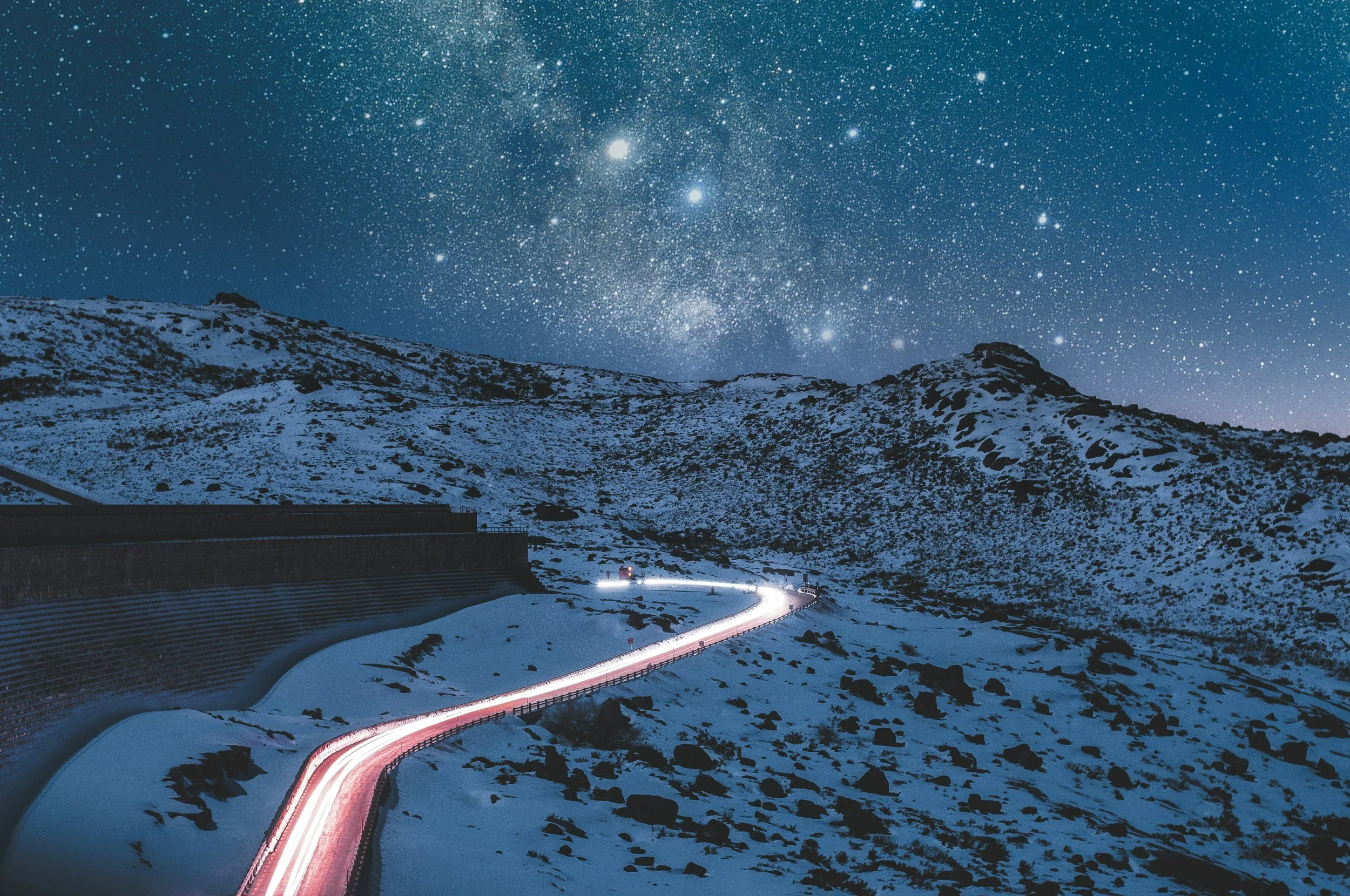 Snow-covered mountain landscape at night with a winding road illuminated by vehicle headlights and the starry sky overhead.