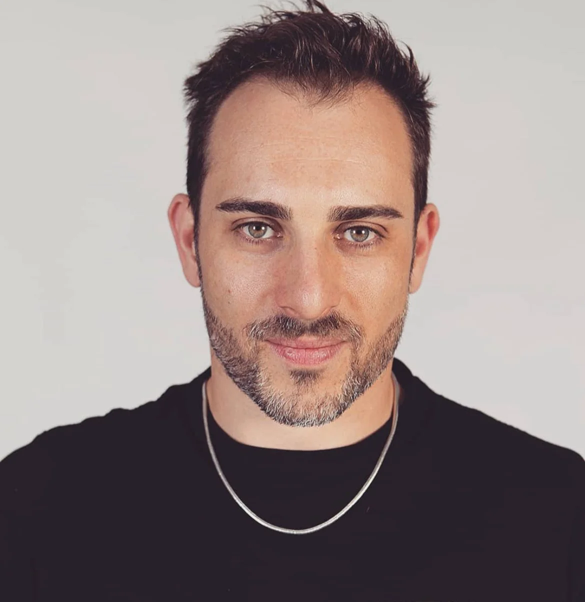Ritratto di un uomo con barba cortissima, capelli castani, maglietta nera e collana argentata, con sfondo bianco.