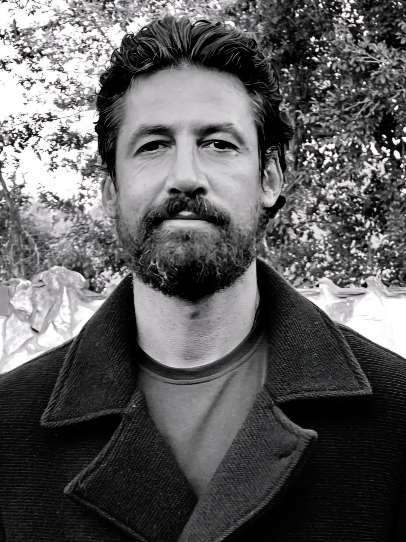 ritratto in bianco e nero di un uomo con barba e capelli spiegazzati, indossa una giacca e un maglione, sullo sfondo ci sono alberi.
