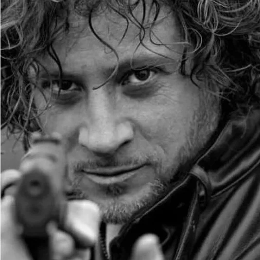 Primo piano di un uomo con capelli ricci e barba che punta una pistola verso la fotocamera.