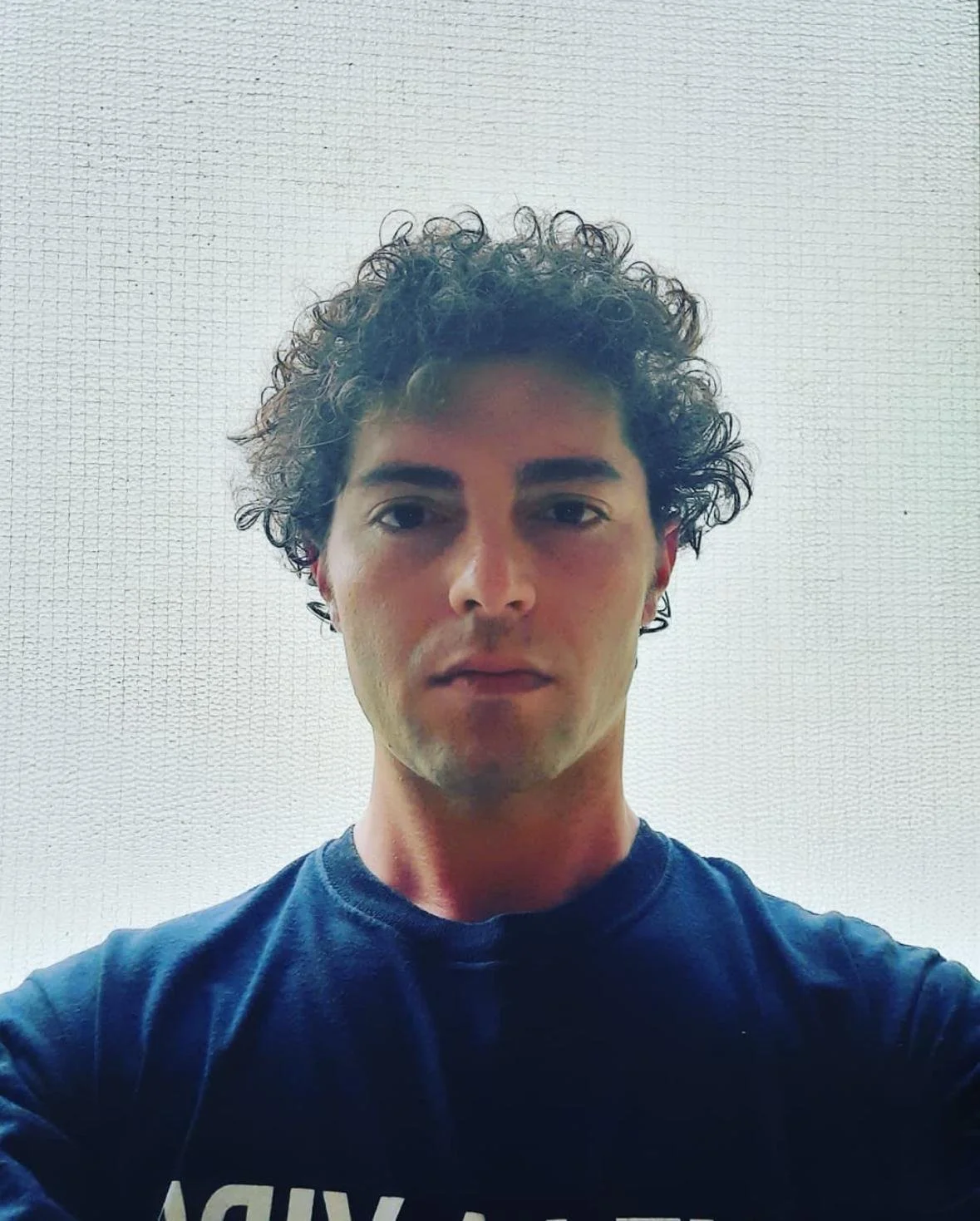 Ragazzo con capelli ricci e scuri, che indossa una maglietta blu, in piedi davanti a un muro bianco.