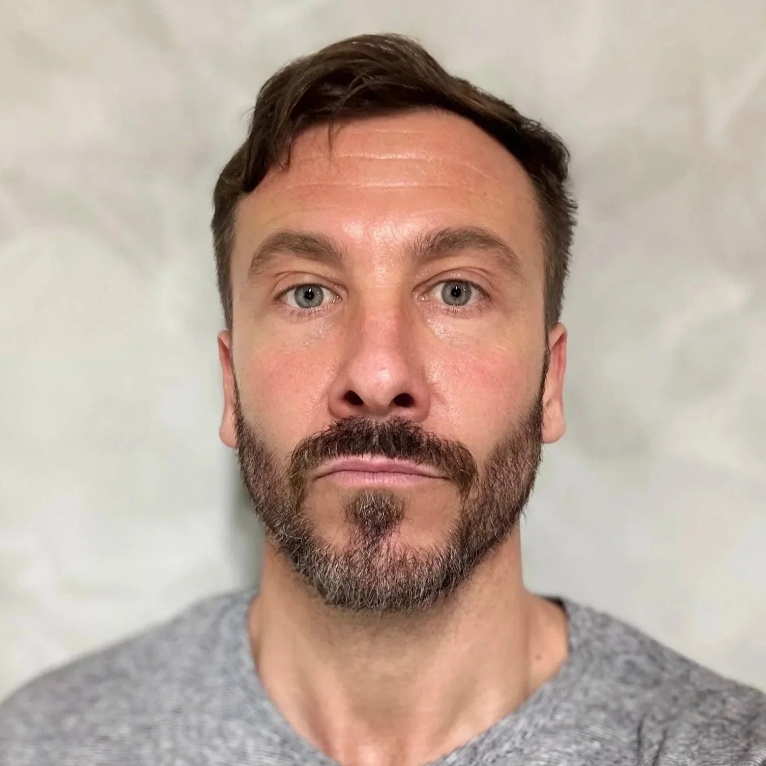 Ritratto di un uomo con barba, cappello corto di capelli castani e maglietta grigia, con sfondo neutro.