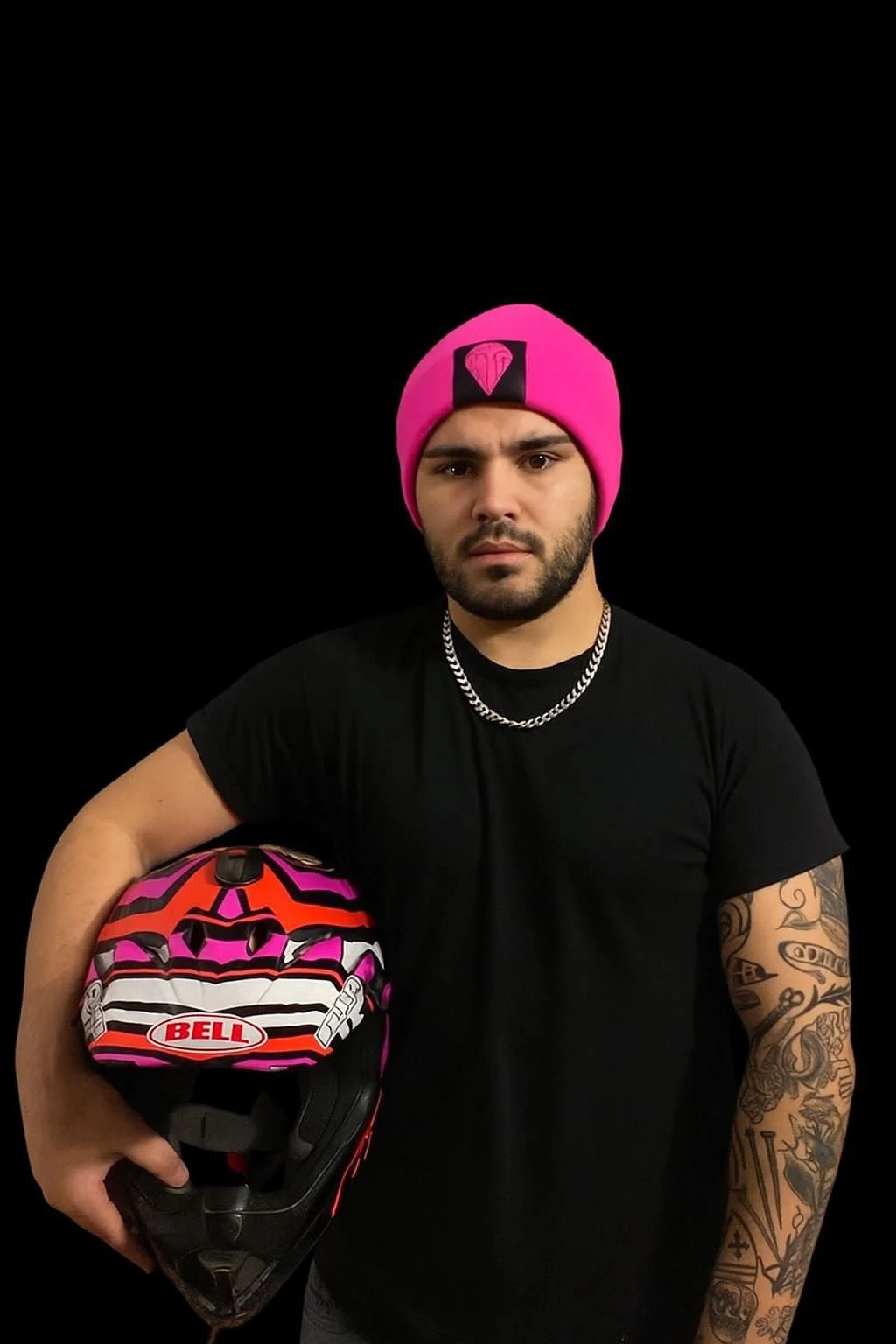 Uomo con barba, tatuaggi sulle braccia, maglietta nera, catena al collo, cappellino rosa con logo, che tiene un casco da moto a tema rosa e nero.