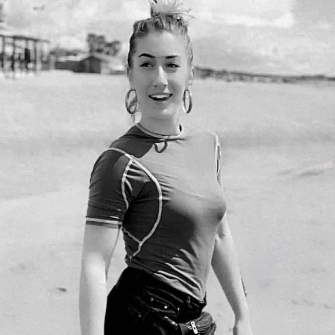 Una donna felice in piedi sulla spiaggia con sguardo verso sinistra, sfondo di mare e strutture di legno, in bianco e nero.