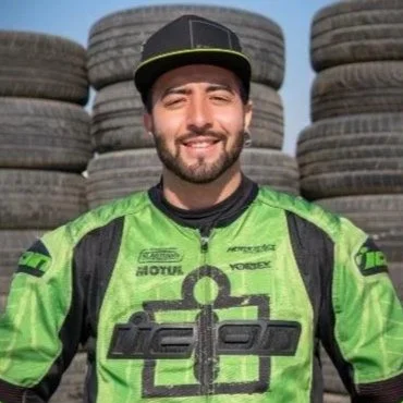 Uomo sorridente in tuta da corsa verde con caschetto nero, davanti a un mucchio di pneumatici