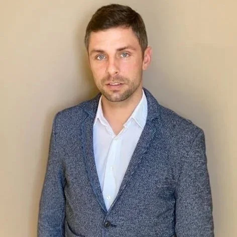 Uomo con capelli corti e barba, indossa un blazer blu e una camicia bianca, posa contro uno sfondo beige.