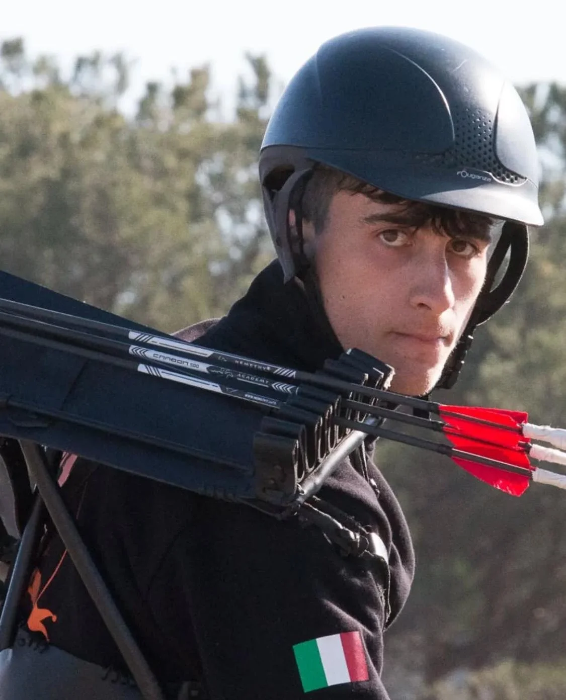 Un giovane atleta con casco e giacca nera, porta frecce da tiro con arco sulle spalle, indossa una maglia con il tricolore italiano.
