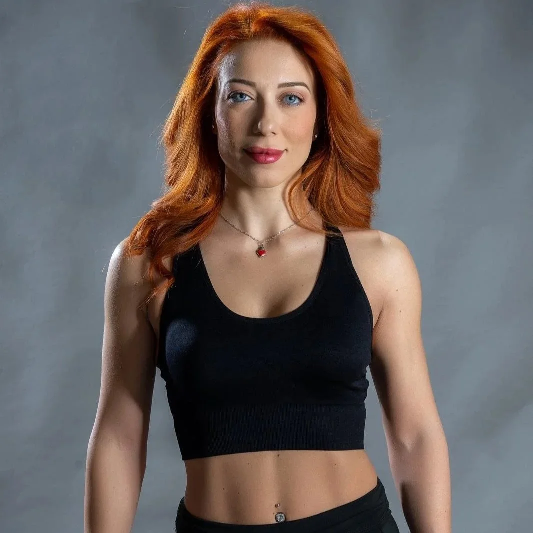 Donna con capelli rossi, abbigliamento sportivo e sorriso, posa di fronte a uno sfondo grigio.