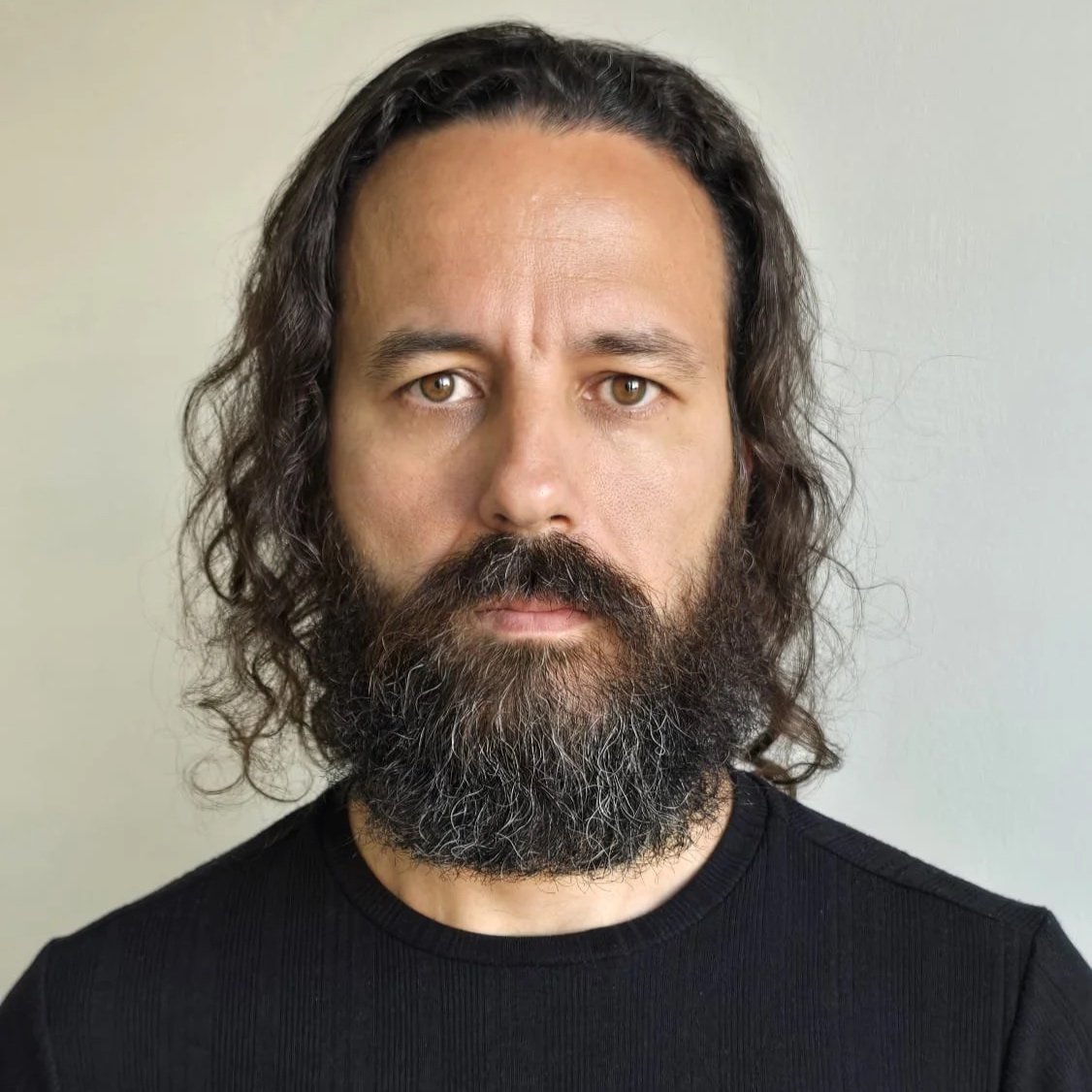 Primo piano di un uomo con capelli ricci, barba folta e occhi marroni, indossa una maglietta nera, sfondo neutro.