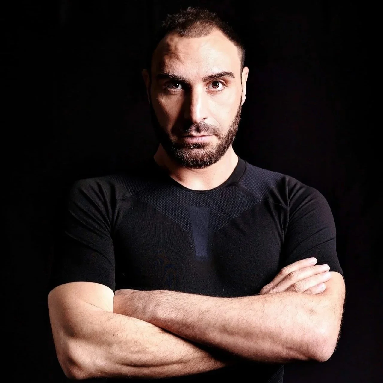 Uomo con barba e capelli corti, sguardo diretto e braccia incrociate, indossa una maglia nera su sfondo nero.