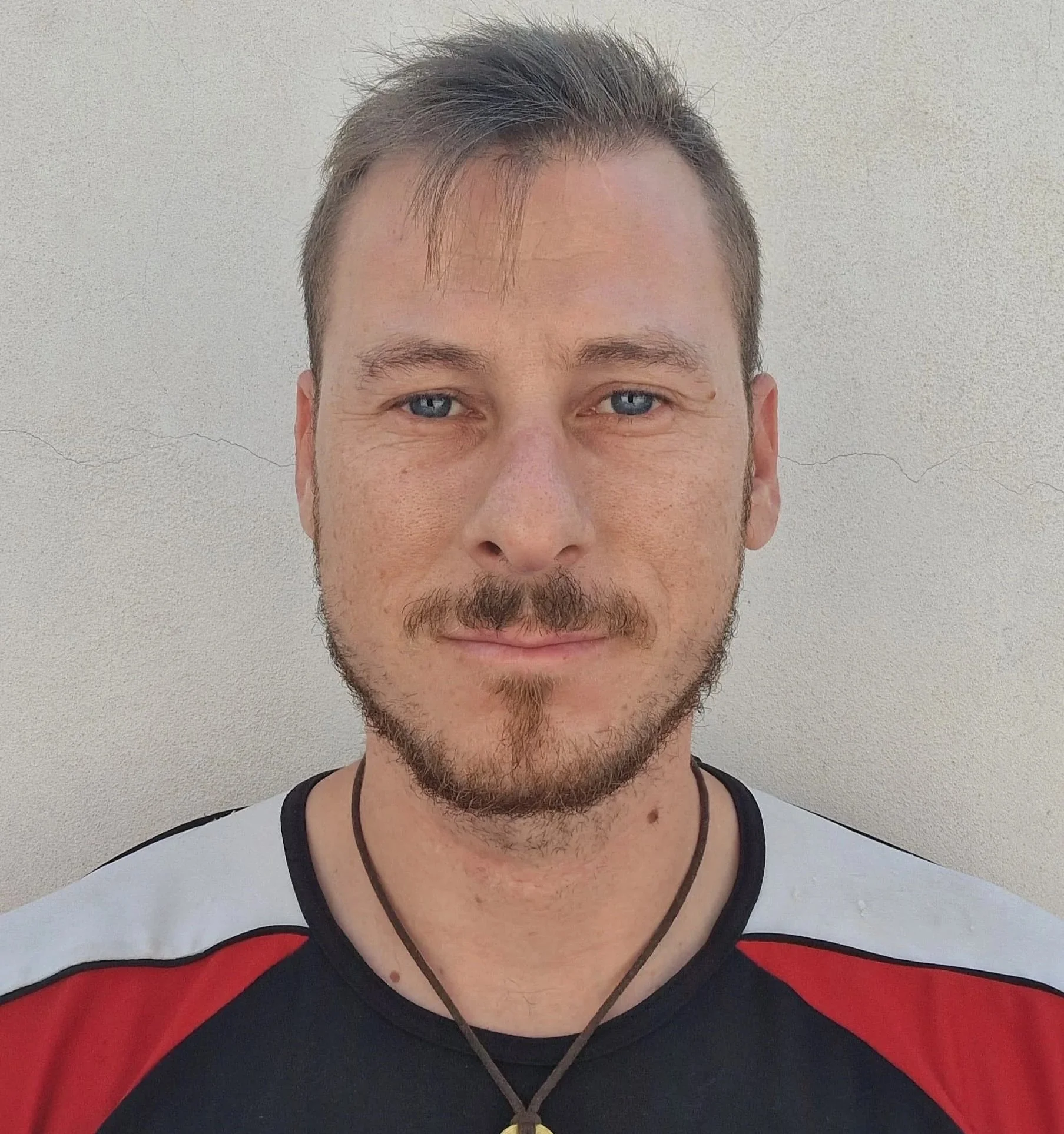 Primo piano di un uomo con capelli corti, barba leggera e occhi azzurri, che indossa una maglietta sportiva a colori nero, rosso e bianco, e una collana con un ciondolo tondo, davanti a una parete beige.