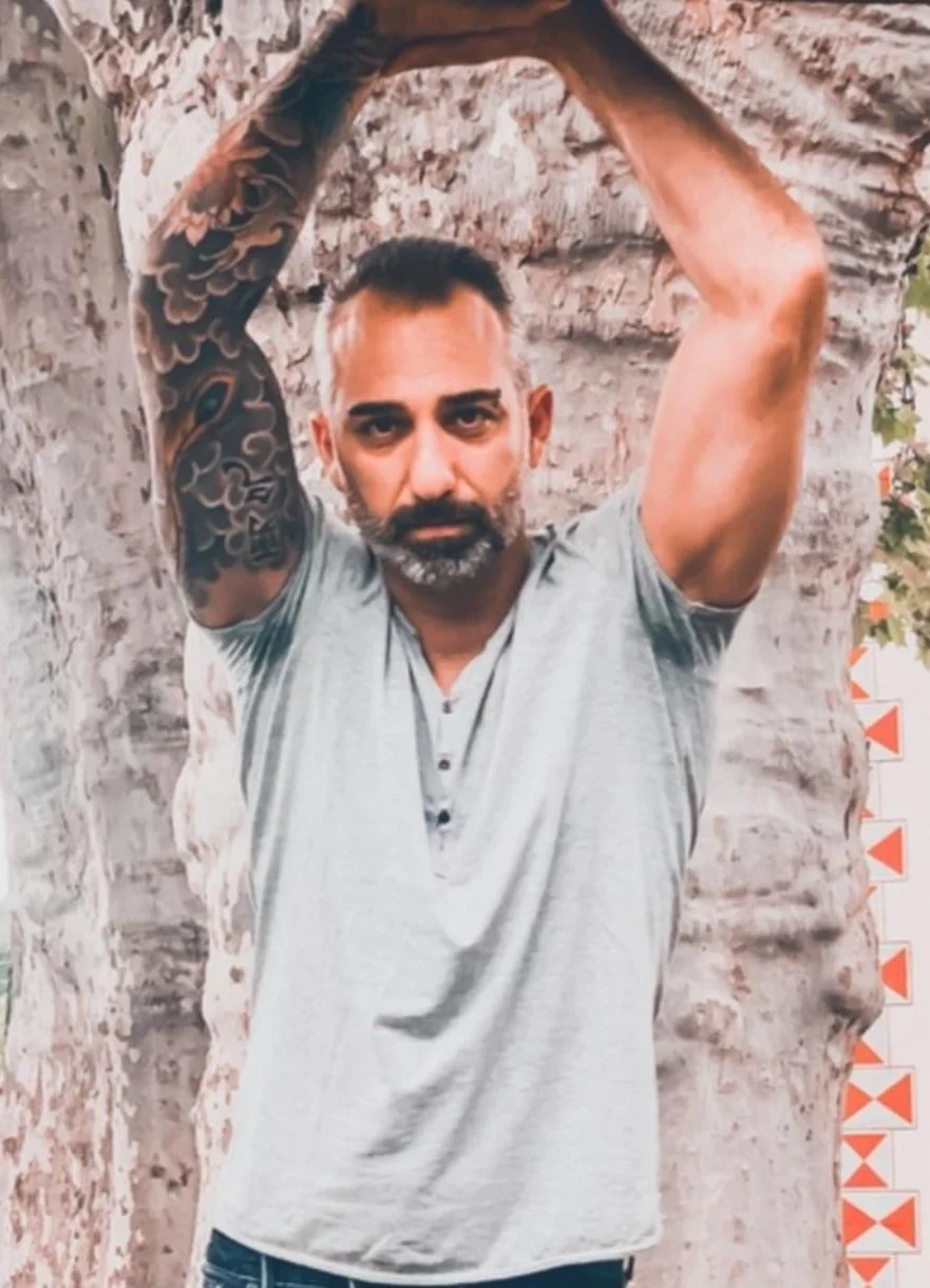Uomo con tatuaggi sulla spalla sinistra, capelli corti, barba, indossa maglietta grigia e si trova davanti a un albero con corteccia ruvida.