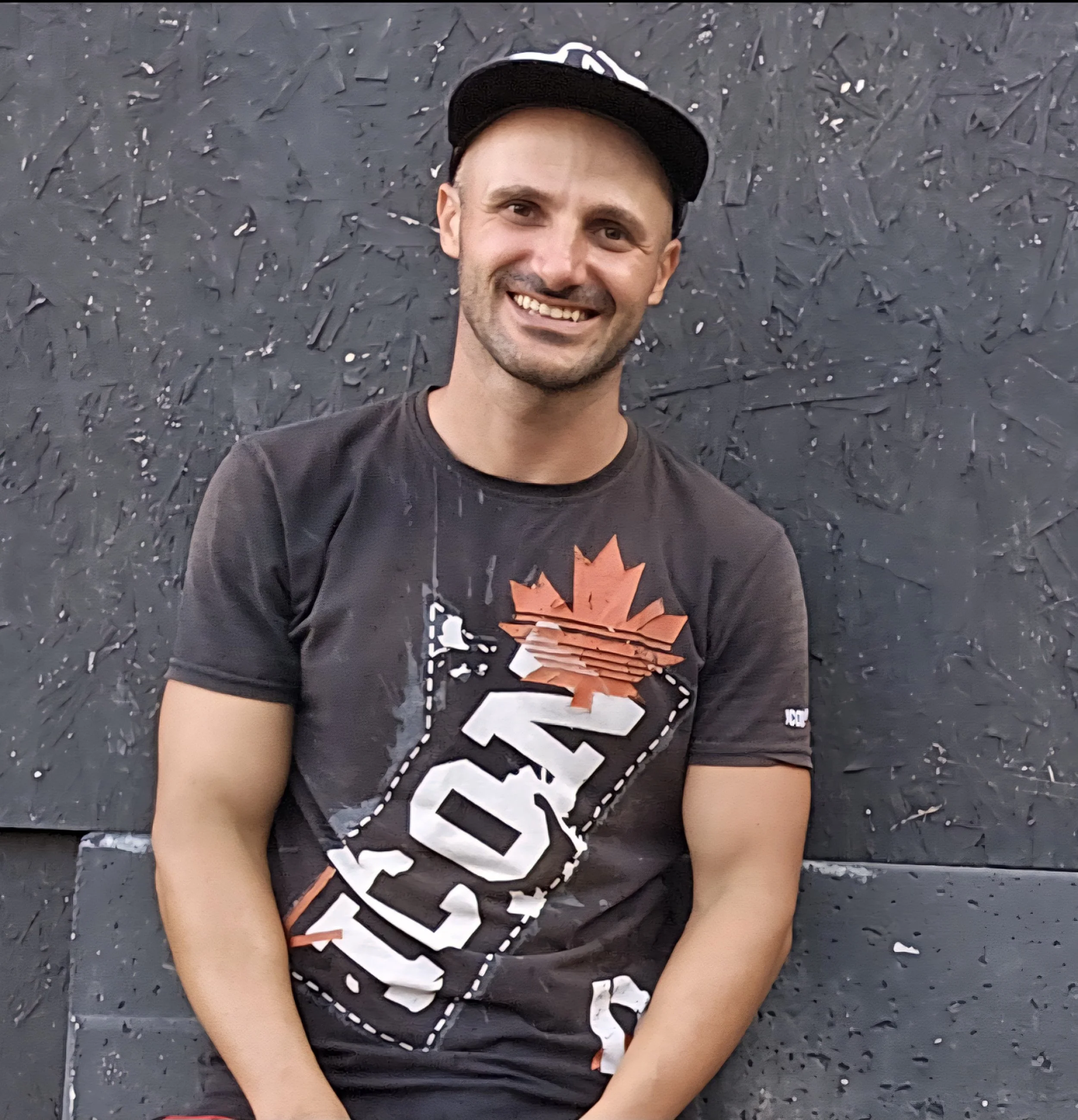 Uomo sorridente con maglietta con logo di Canadian Tire e cappellino nero, mentre posa con braccia incrociate davanti a una parete nera.