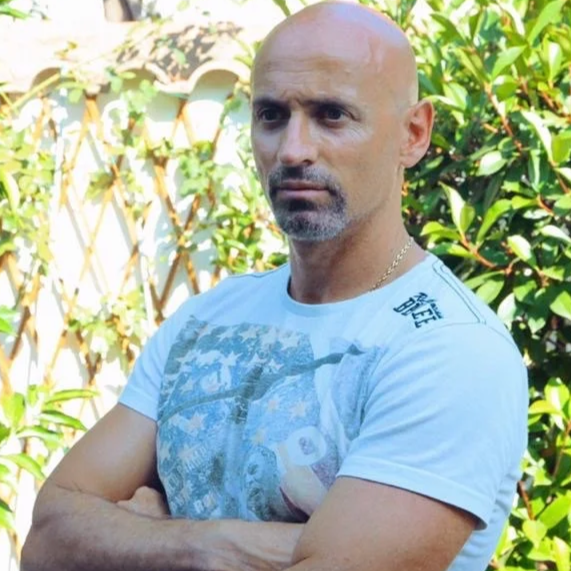 Uomo con cappello a capanna, braccia incrociate, di fronte a una recinzione coperta di piante verdi.