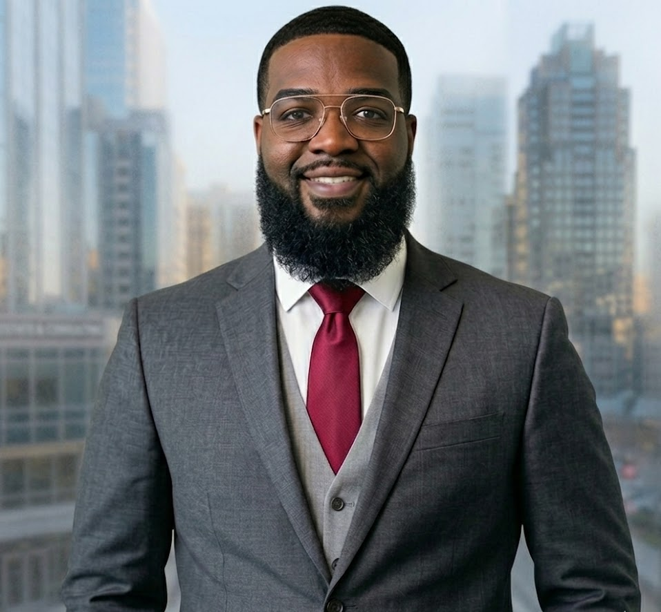 A smiling African American man in a gray suit with a red tie, standing in front of a city skyline with tall buildings.