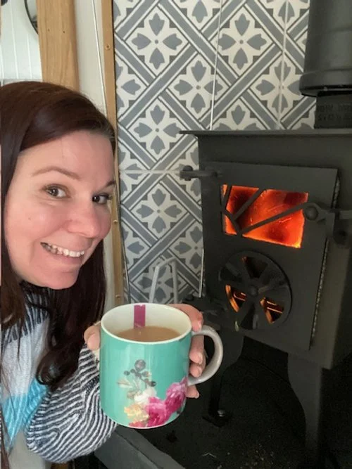 A woman smiling and holding a mug of coffee or tea in front of a black wood stove with glowing fire inside. The background has patterned blue and white tiles.