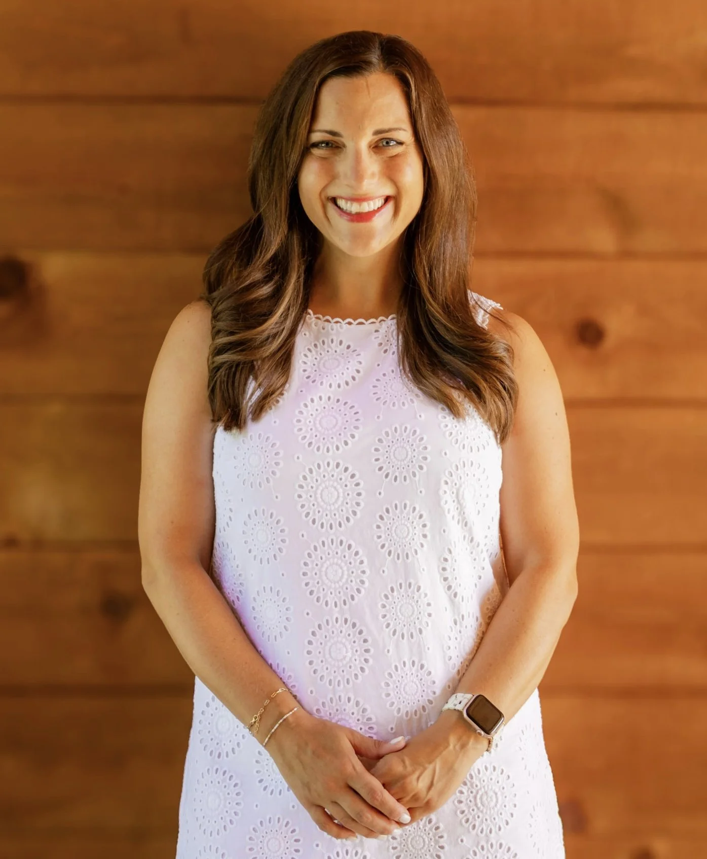 A woman with long brown hair smiling and standing against a wooden background, wearing a sleeveless white dress with eyelet embroidery, a watch, and jewelry.