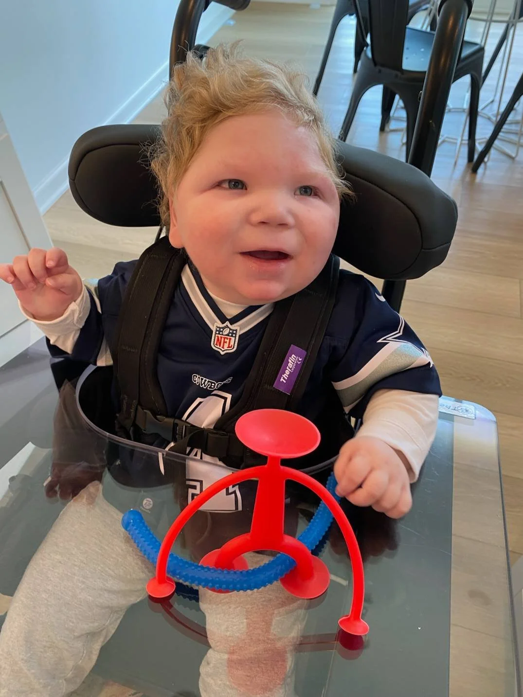 A young child with curly blonde hair sitting in a high chair at a table, wearing a Dallas Cowboys jersey, with a red and blue sensory toy in front of him on the table.