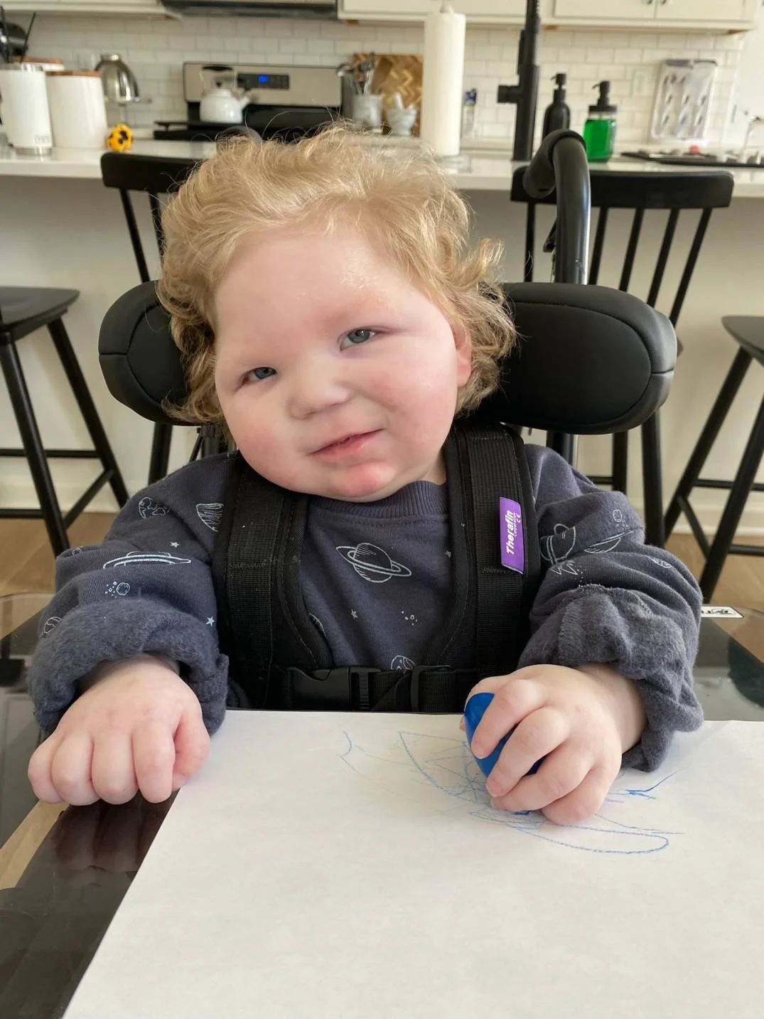 Young child with curly red hair sitting at a table, holding a blue crayon, with a piece of paper and colorful scribbles in front. The child wears a space-themed shirt and a black harness, in a kitchen or dining area.