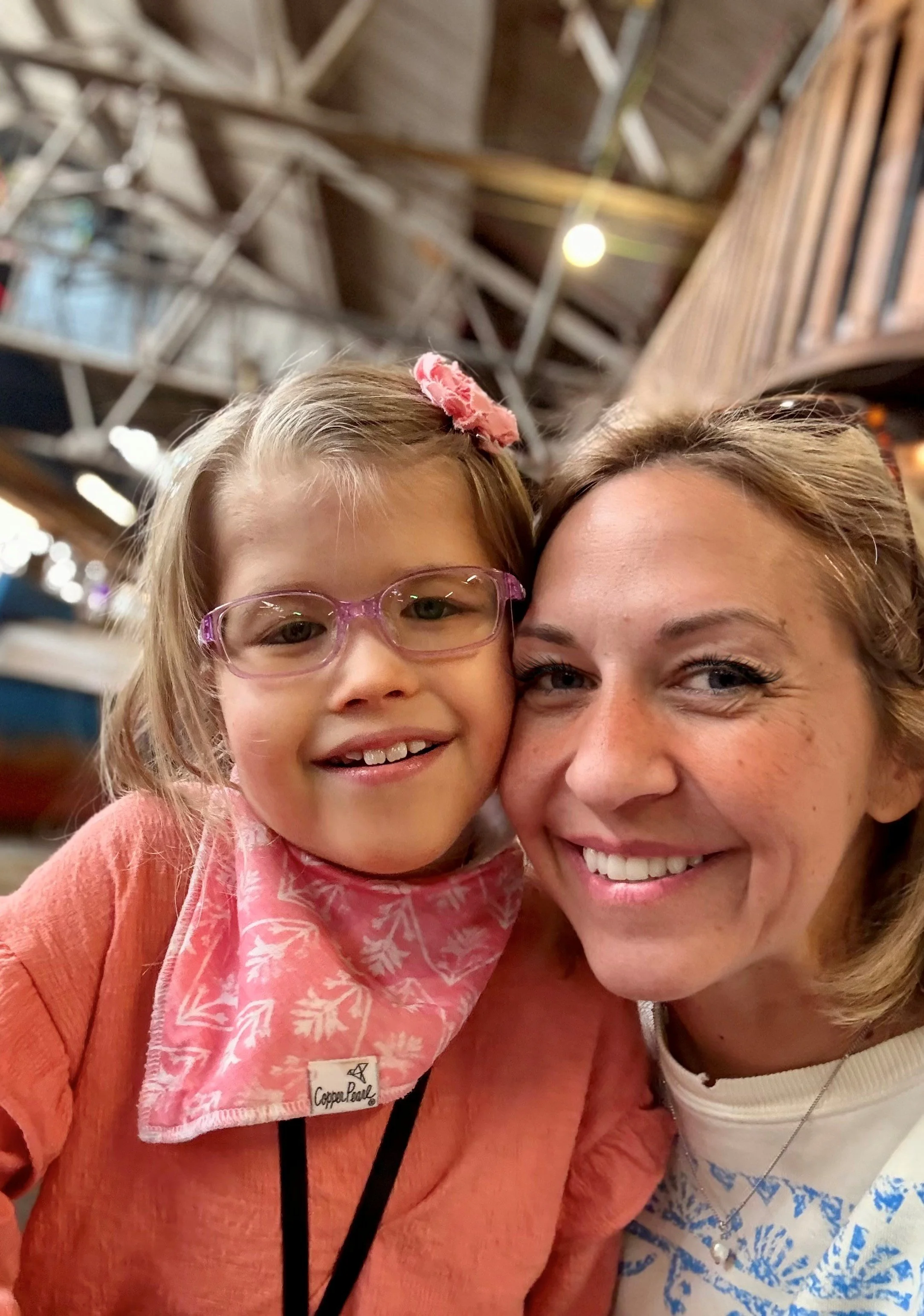 A woman and a young girl taking a selfie together in a rustic indoor setting. The girl is wearing pink glasses, a pink bandana, and a peach-colored shirt. The woman has short blonde hair, is smiling, and wearing a white shirt with blue patterns and a necklace.