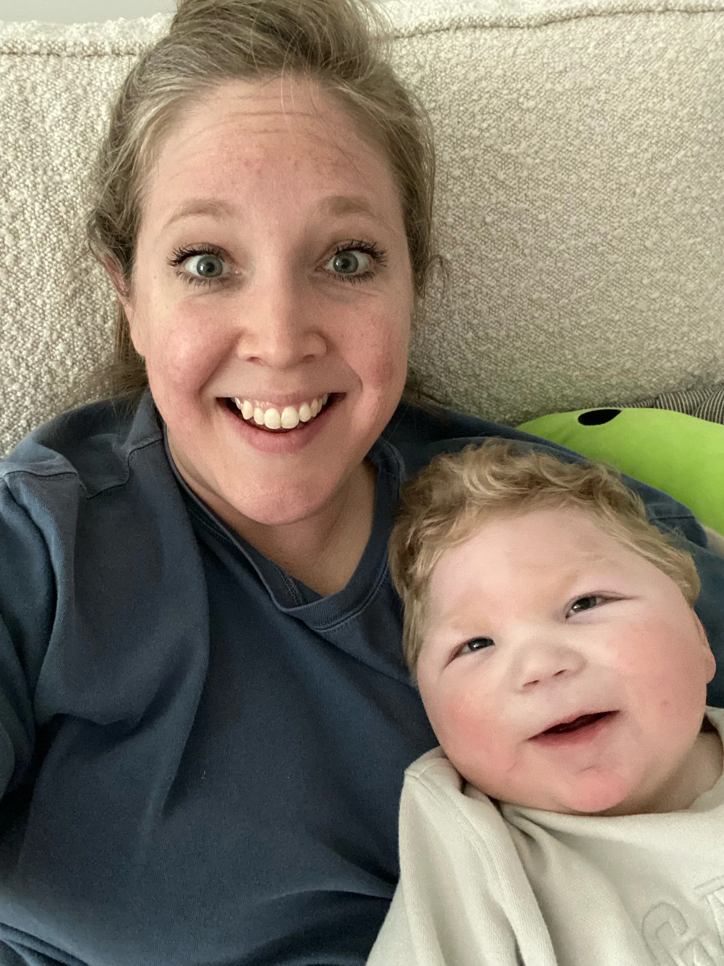 A woman with light skin and blonde hair smiling, sitting with a young child with red curly hair and chubby cheeks, both looking at the camera, on a beige couch with a green ball in the background.