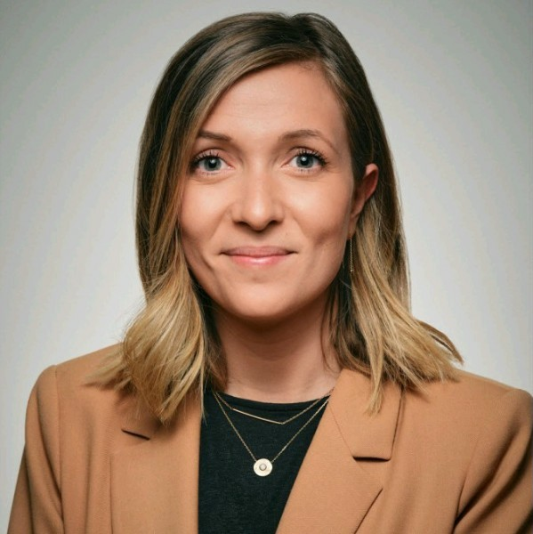 Une femme souriante avec des cheveux mi-longs, habillée d'une veste beige et portant des bijoux, posant contre un fond neutre.