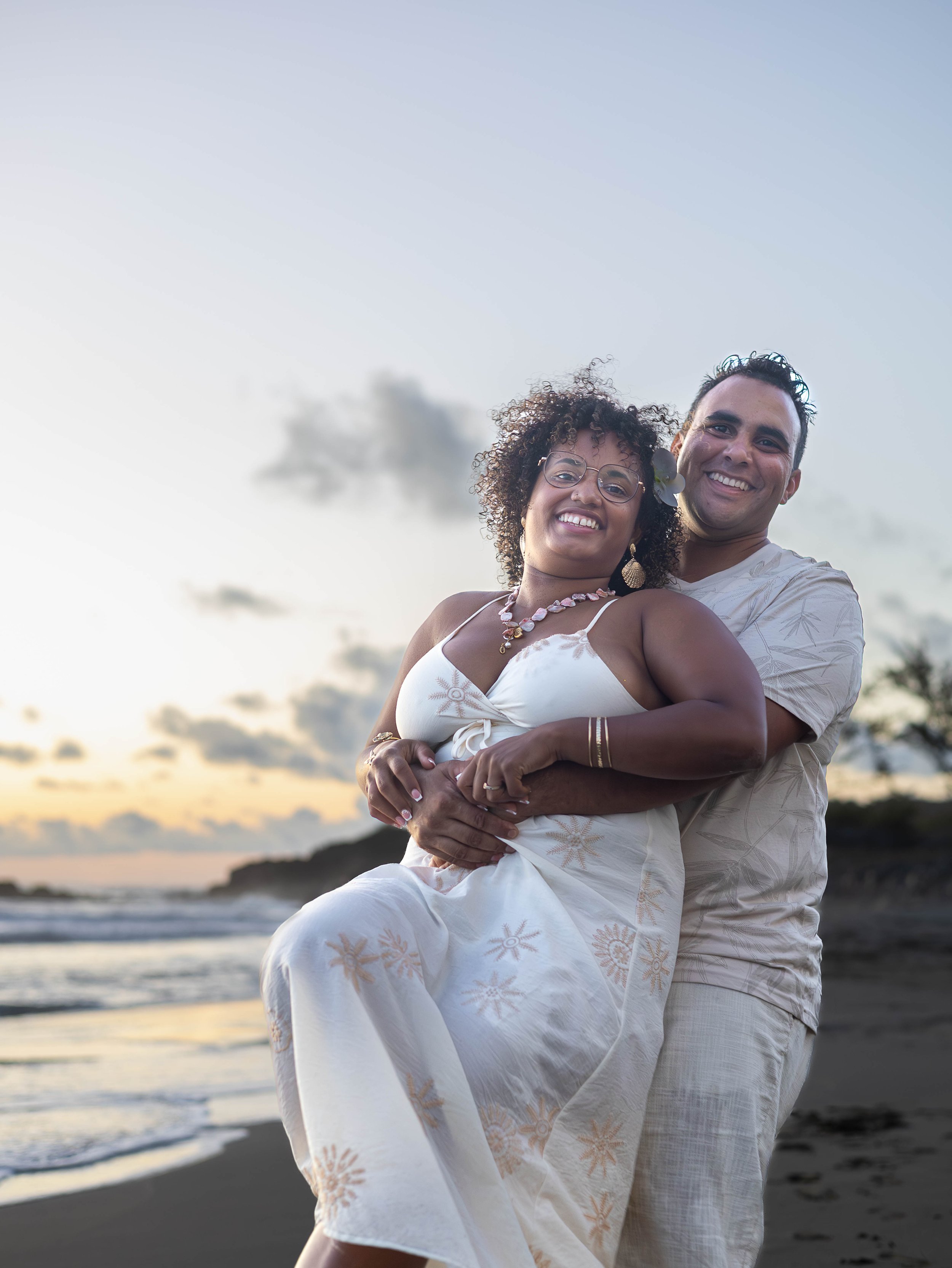 Un couple souriant lors d'une séance photo sur la plage au coucher du soleil, portant des vêtements légers et colorés.