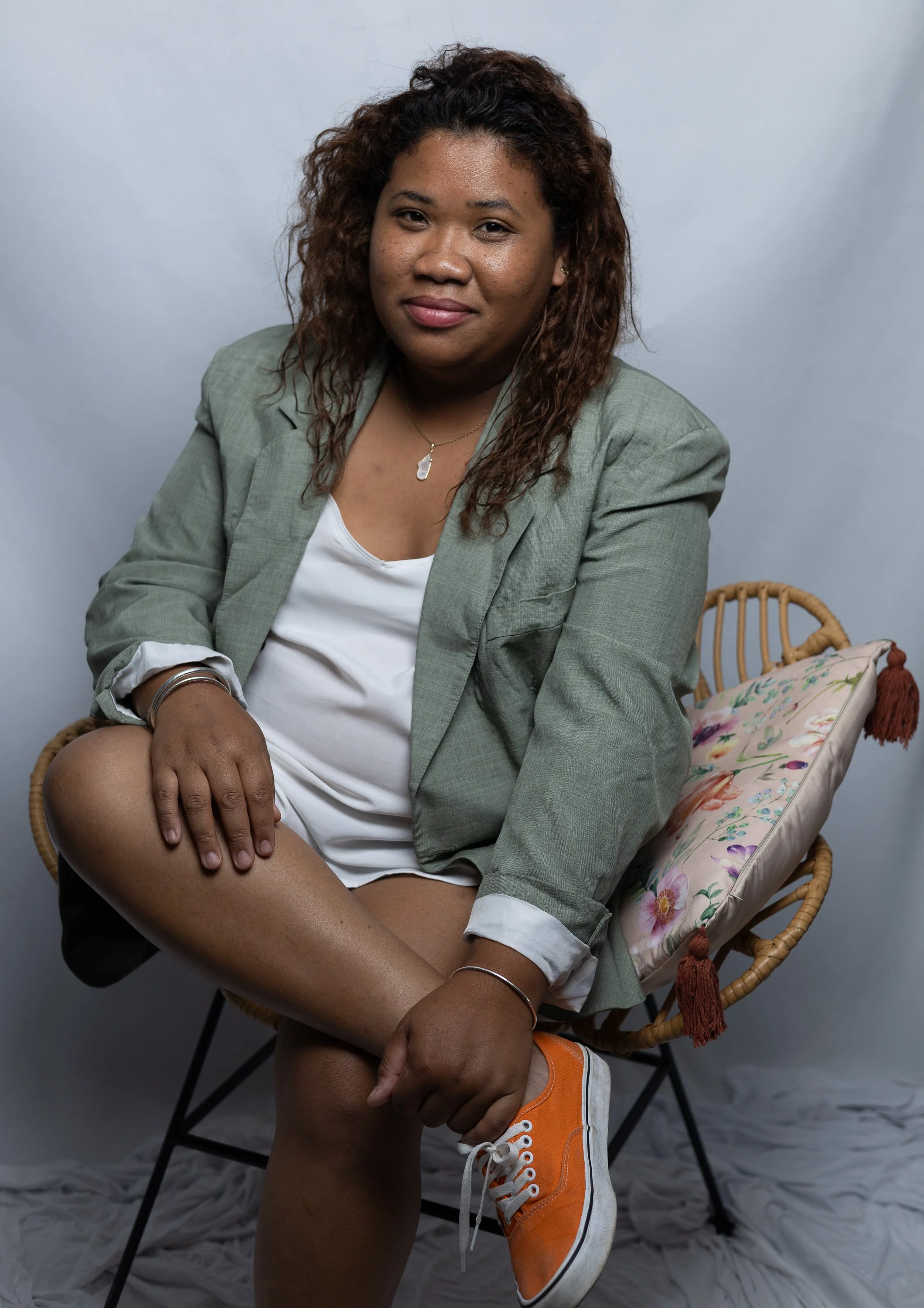 Une femme assise sur une chaise en osier, portant une veste verte, une chemise blanche et une paire de baskets orange. Elle a des cheveux bouclés et foncés, et regarde vers l'objectif avec un léger sourire.