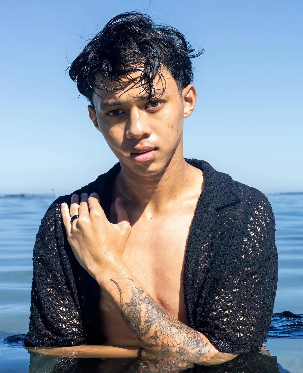 Jeune homme nu jusqu'au ventre, portant une veste en crochet noir, dans l'eau, avec un tatouage à l'avant-bras, regardant l'objectif, fond de mer et ciel bleu.