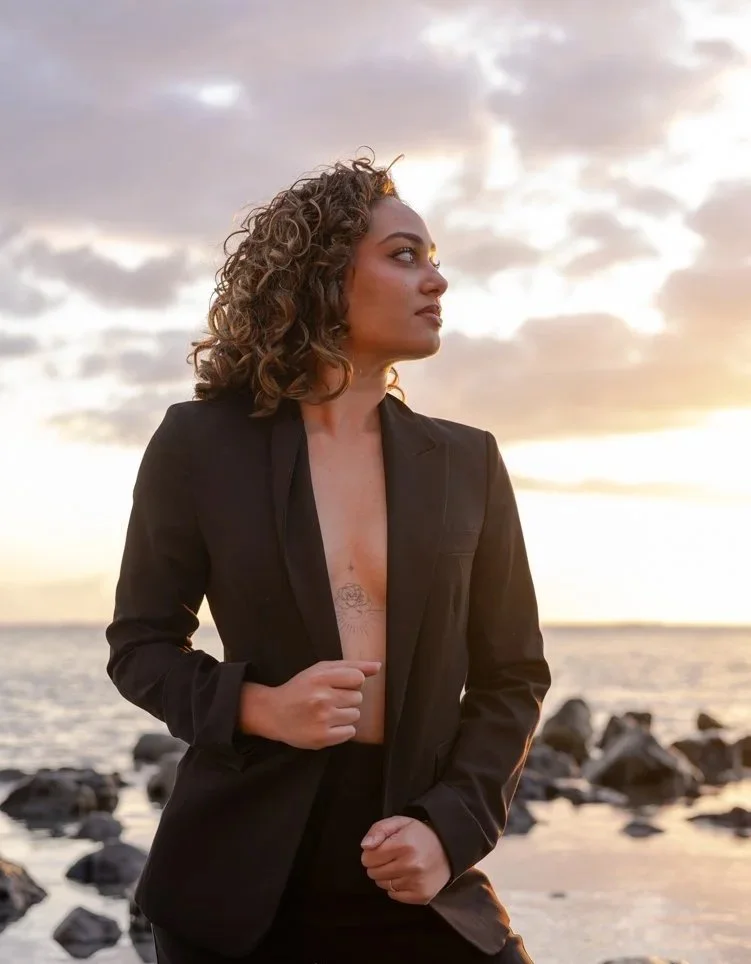 Femme avec des cheveux bouclés portant un costume noir, debout sur une plage au coucher du soleil.