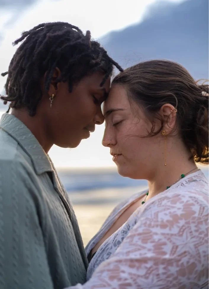 Deux femmes se tiennent proches avec leurs fronts touchant, dans un cadre naturel avec la mer en arrière-plan, lors du coucher de soleil.