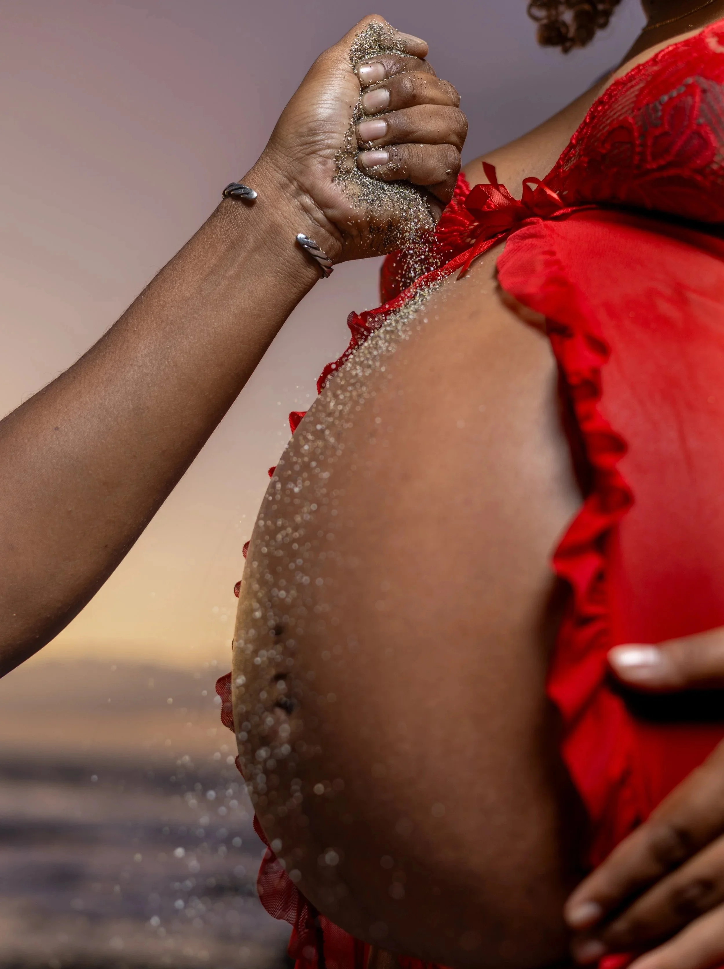 Une personne en robe rouge avec des détails en dentelle tient ses mains jointes devant son épaule, avec des grains de sable ou de brillant tombant ou posé dessus.